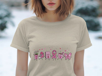 Tan t-shirt with a cartoon line of pink breast cancer awareness ribbon, flower pot, shoe, notebook, mug, and butterfly.