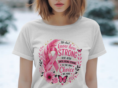 White t-shirt with pink ribbon, flowers, feathers, and butterflies. Text reads: "We don't know how strong we are until being strong is the only choice we have."