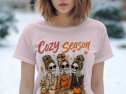 Pink t-shirt with "Cozy Season" text and three skeleton friends holding drinks and pumpkins.