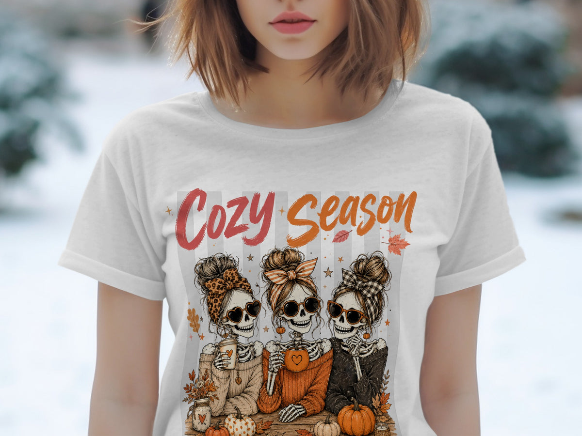 Three skeleton women with sunglasses and buns sit with coffee mugs and pumpkins, text reads "Cozy Season".